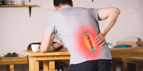 Man in a workshop leaning on table, holding his lower back in pain, depicted with red flare and spine graphic.