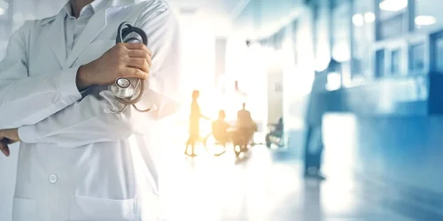 Doctor in white coat holding a stethoscope with blurred patients in background in a hospital.