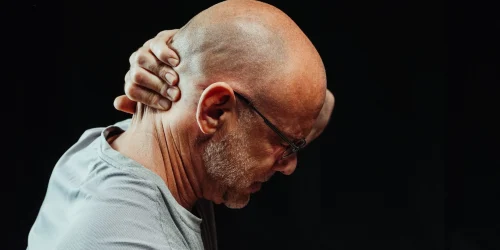Bald man in a gray shirt grasping his head in pain or distress against a black background.