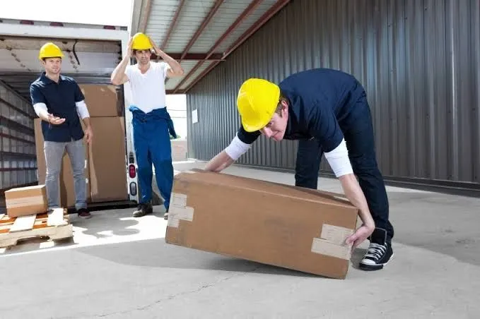 Three workers in hard hats, one lifting a heavy box in a warehouse.