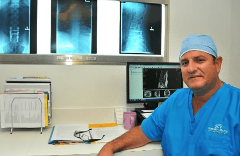 Doctor in blue scrubs reviewing X-rays in an office with a computer and files.