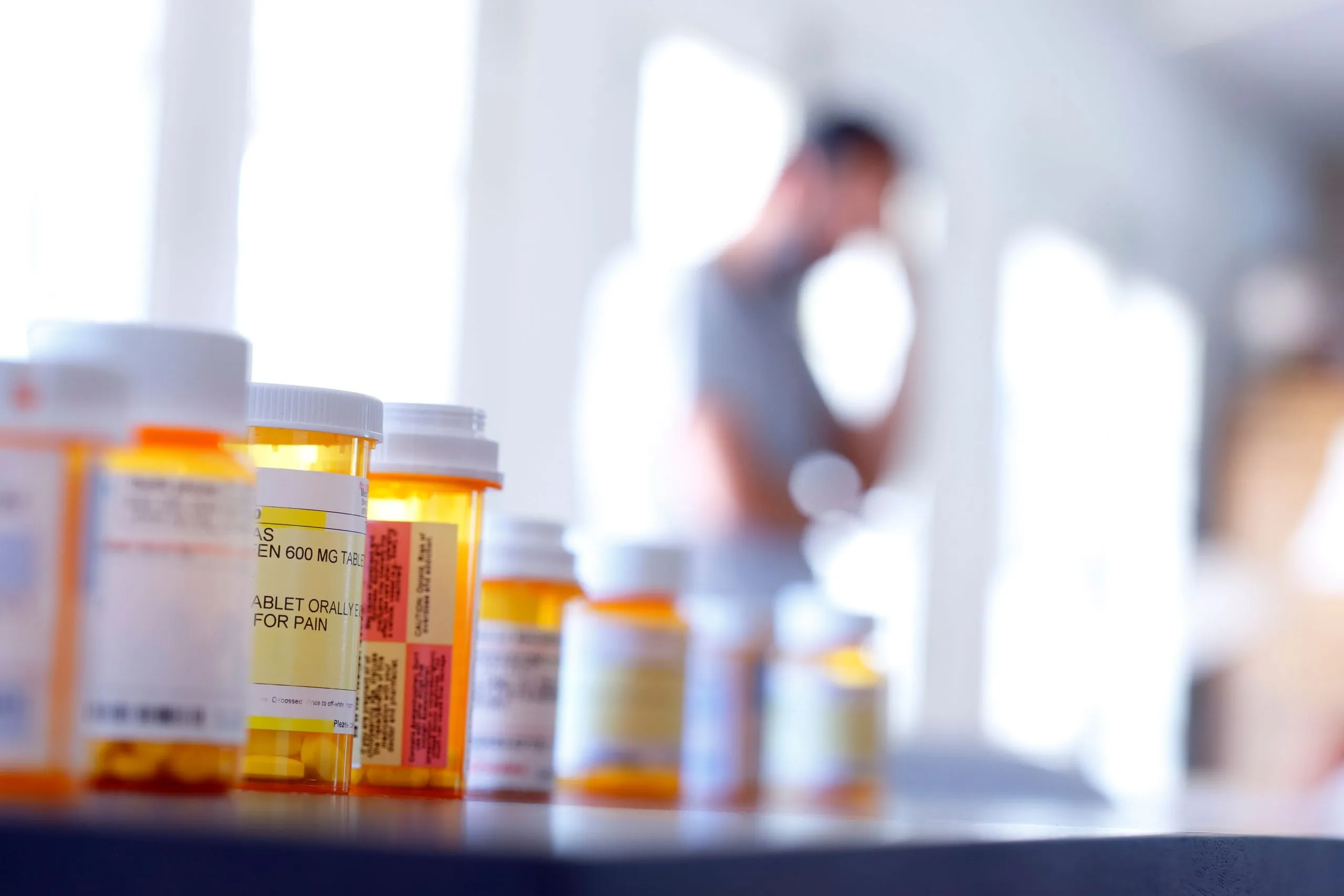 Bottles of prescription pain medication in focus with a blurred man in the background, suggestive of the increased use of opioids and pain medications.