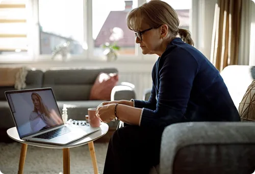 A woman sitting in a living room communicates via video call on a laptop.