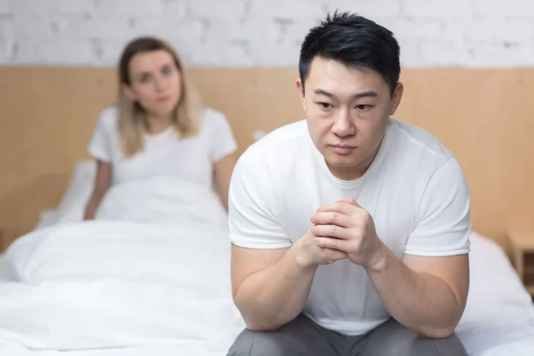 A man sits on the edge of a bed looking pensive, while a woman sits behind him, slightly blurred.