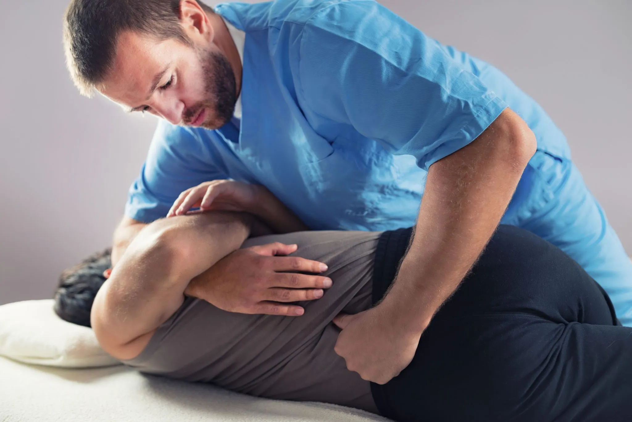 A physical therapist in blue scrubs is performing manual therapy on a male patient lying prone on a therapy table, suggesting a scenario where physical therapy might not work if not properly tailored to the patient's condition.