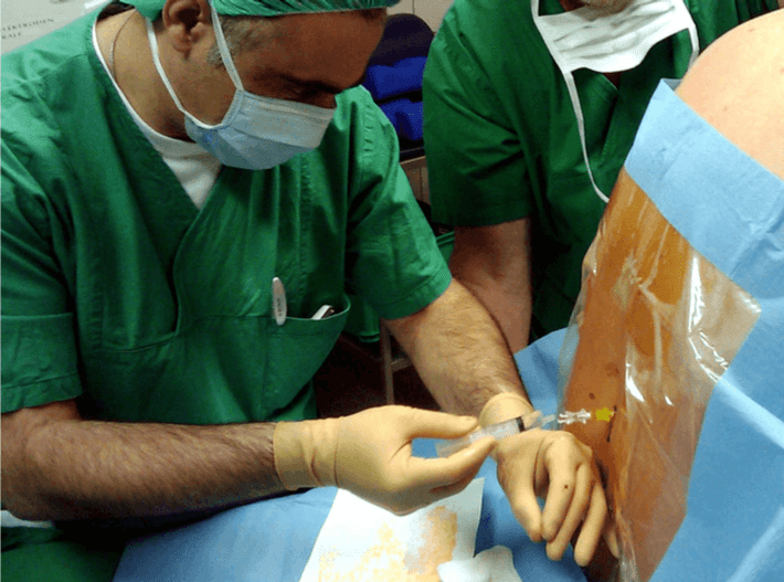 A medical professional in green scrubs administers an injection into a patient's back.
