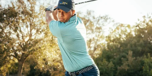 Golfer in mid-swing with a driver, wearing a striped shirt and cap on a sunny day.